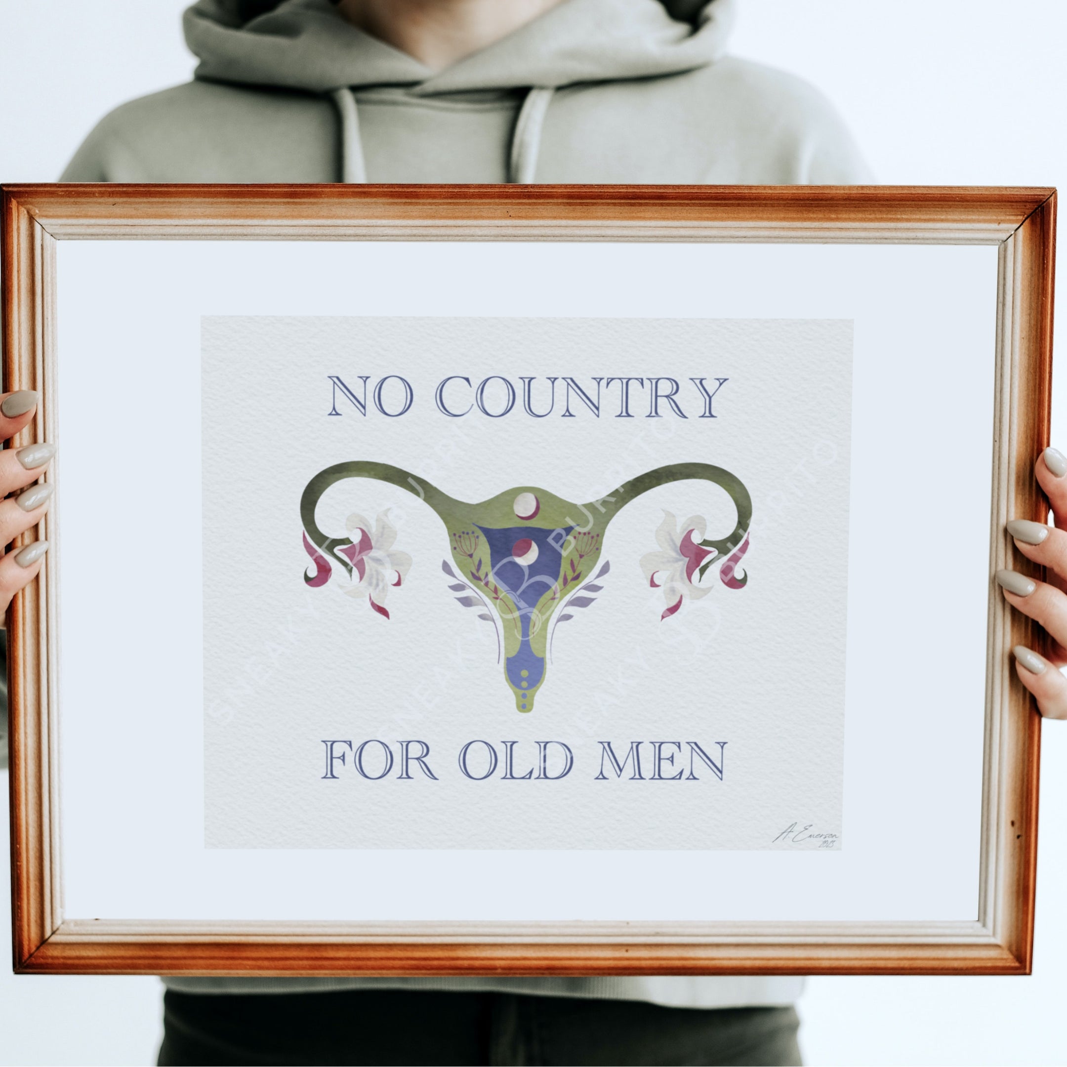 A person in a light gray hoodie holds a framed art print featuring a stylized uterus illustration with the text 'NO COUNTRY FOR OLD MEN' in blue serif lettering. The print has a white mat and a warm brown wooden frame.