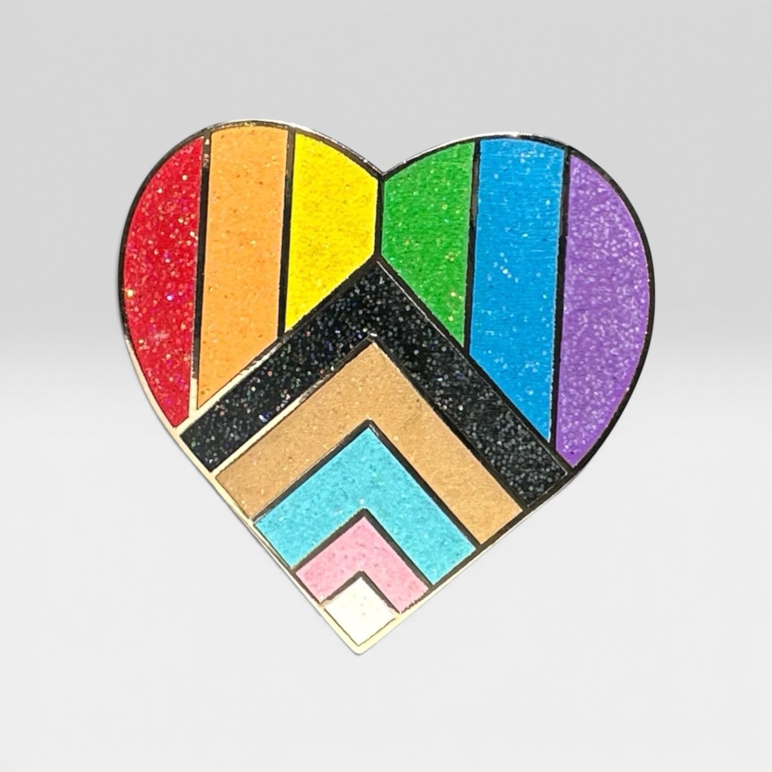 A heart-shaped enamel pin featuring the Progress Pride flag design with glittery stripes and chevron patterns in red, orange, yellow, green, blue, purple, black, brown, light blue, pink, and white, on a light gray background.