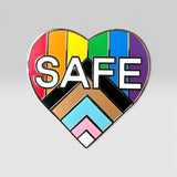A heart-shaped enamel pin featuring the word 'SAFE' in bold white letters over a rainbow-striped background and a Progress Pride flag motif at the bottom, shown on a light gray background.