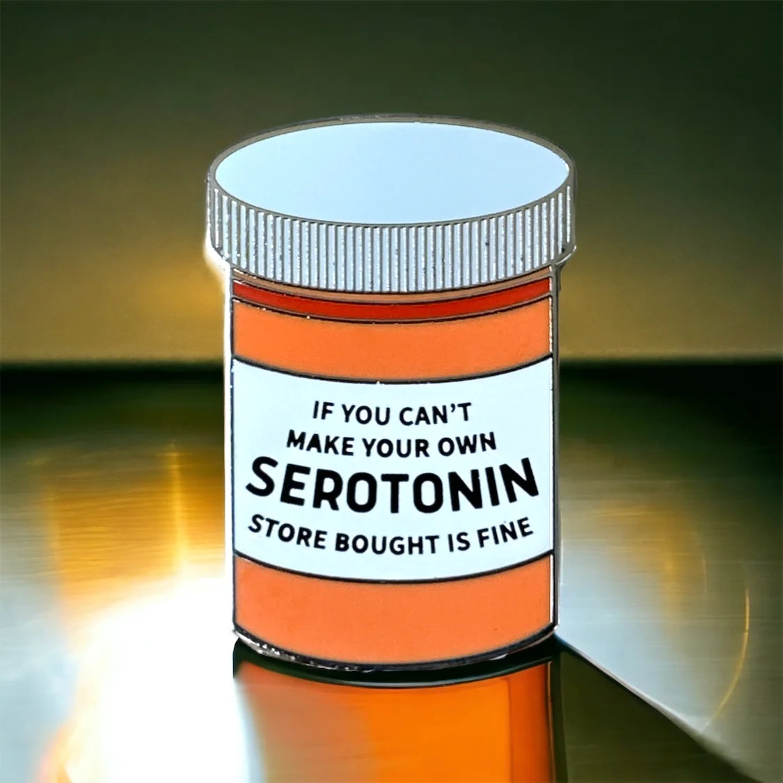 A pill bottle-shaped product with an orange body, white label, and white cap, shown on a reflective dark surface. The label reads, 'IF YOU CAN'T MAKE YOUR OWN SEROTONIN STORE BOUGHT IS FINE.
