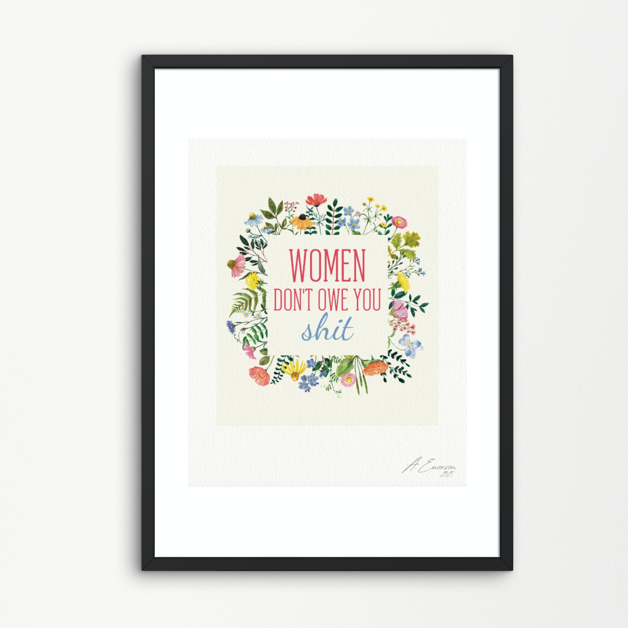 A framed typographic art print featuring a floral wreath around the text 'WOMEN DON'T OWE YOU shit' in pink, red, and blue lettering. The print has a cream background, is mounted with a wide white mat, and is enclosed in a slim black frame against a light gray wall.